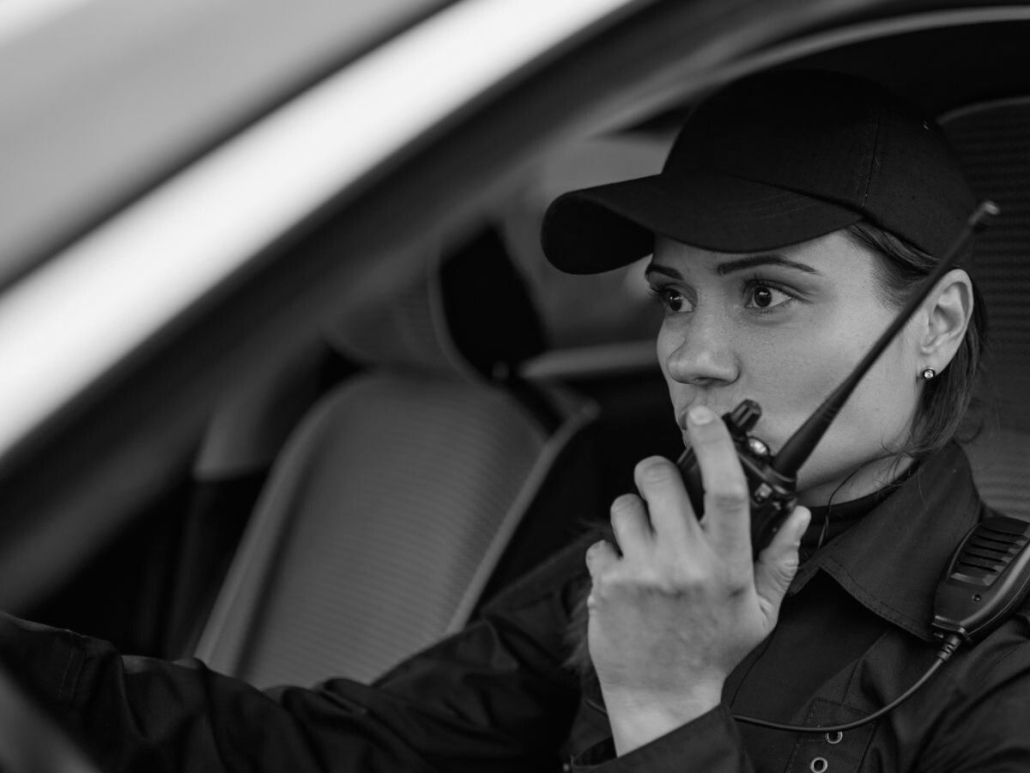 Female security officer in patrol vehicle using two-way radio during mobile security patrol.