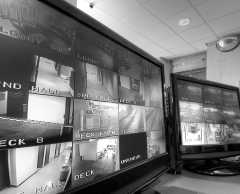 Security monitoring room with multiple CCTV screens displaying live camera views of business entrances, hallways and external areas.