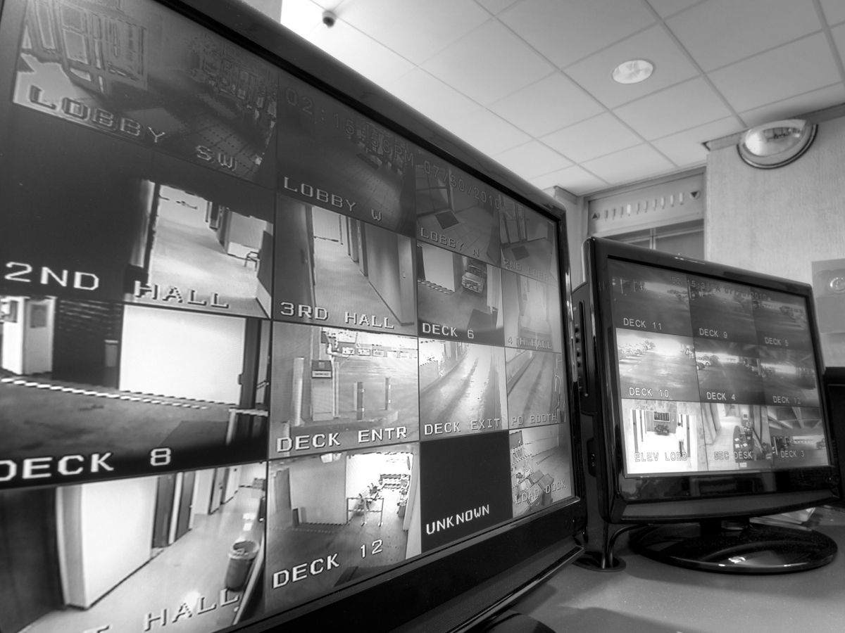 Security monitoring room with multiple CCTV screens displaying live camera views of business entrances, hallways and external areas.