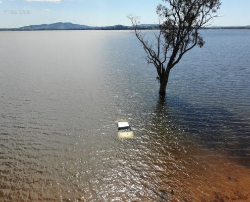 Aerial image by PGC Aerial Gallery showing a submerged vehicle near a tree in floodwater at a regional lake.